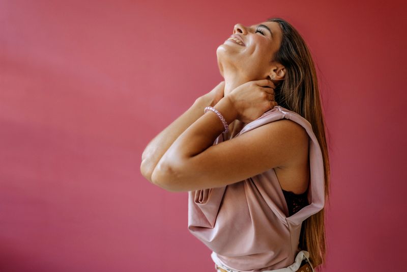A young woman is smiling while standing in a studio with a colorful background. She is gently touching her neck, expressing happiness and confidence. The scene captures a light moment.
