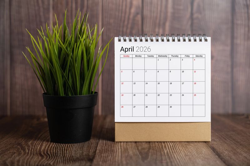 April 2026 desk calendar displayed next to a small potted green plant on a rustic wooden table background.