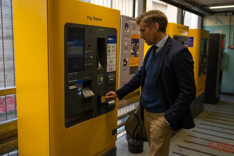 Man in parking garage using credit card at yellow self-service pay station, side view, cashless automated payment, everyday urban routine