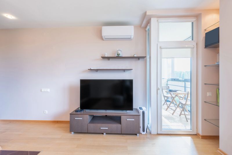 Bright and cozy living room featuring a flat-screen TV, modern furniture, and a balcony door allowing abundant natural light, creating a comfortable and inviting space