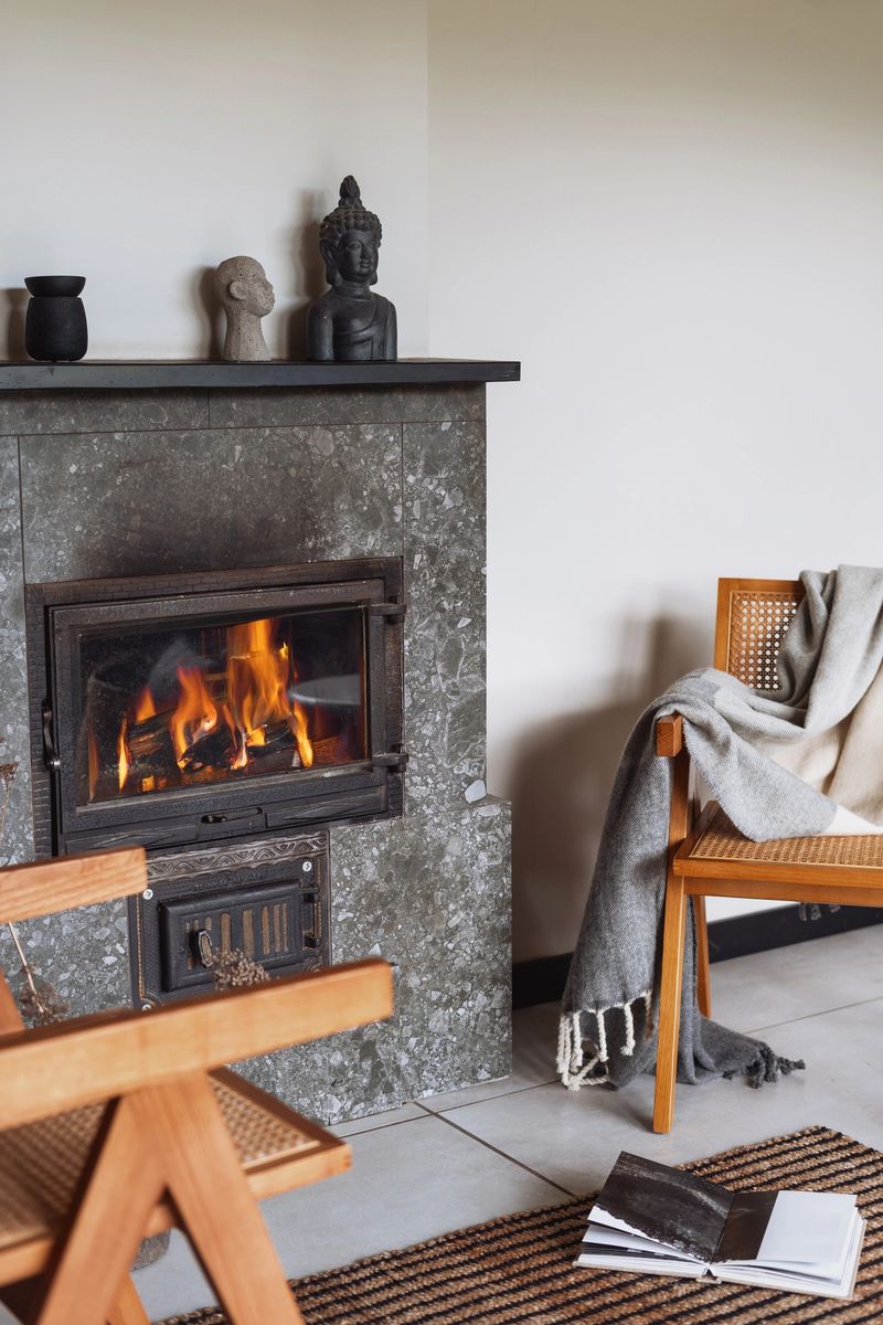 Cozy living room features a modern fireplace, wooden chairs, warm textiles creating comfortable space perfect for relaxing during cold autumn and winter nights. Plaid on armchair close to fire place