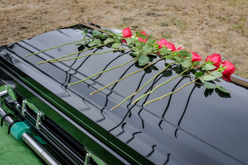 A line of red roses rests gently on a polished casket, their reflections echoing a quiet final goodbye.