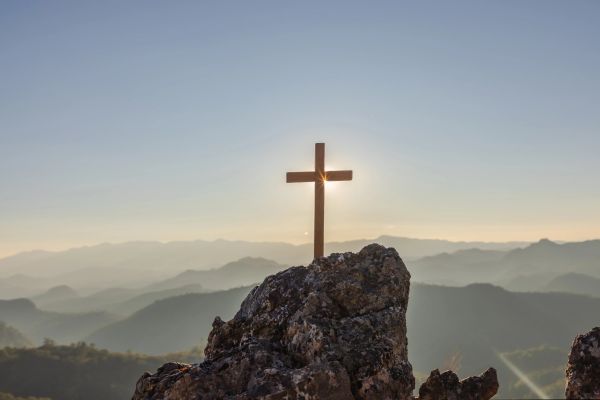 Wooden cross on a rock with sun shining behind it.