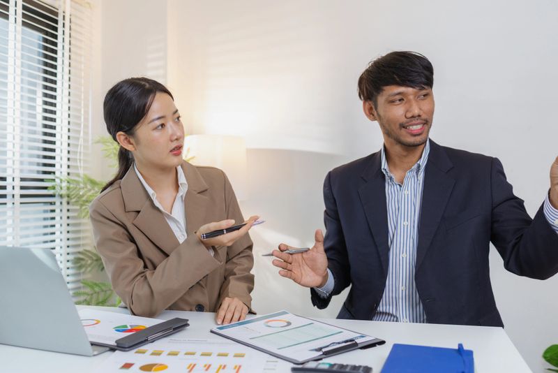 Two business professionals are discussing financial charts and reports in a modern office. They collaborate, share ideas, and analyze data together, highlighting teamwork and strategy.