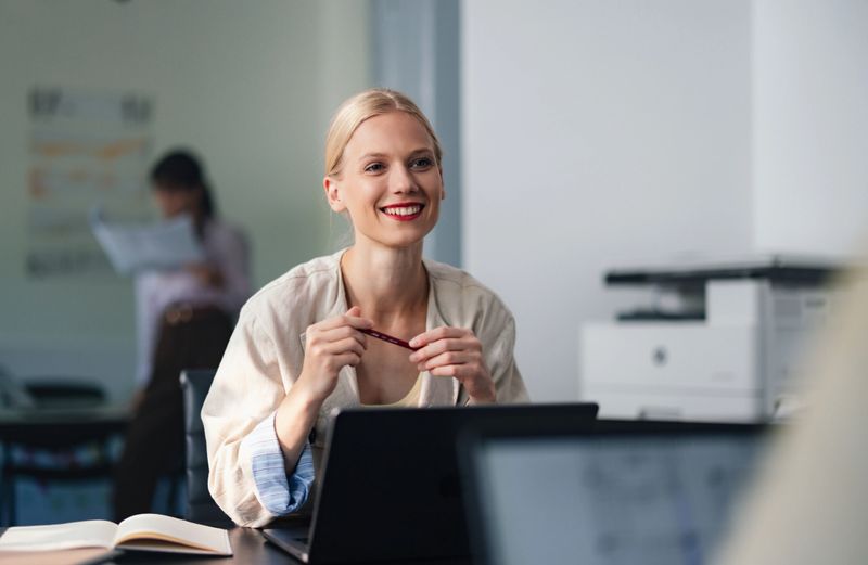 A cheerful professional woman sits at a desk with a laptop, smiling while holding a pen in a bright, modern office. Background shows a blurred coworker and office equipment, conveying productivity and collaboration.