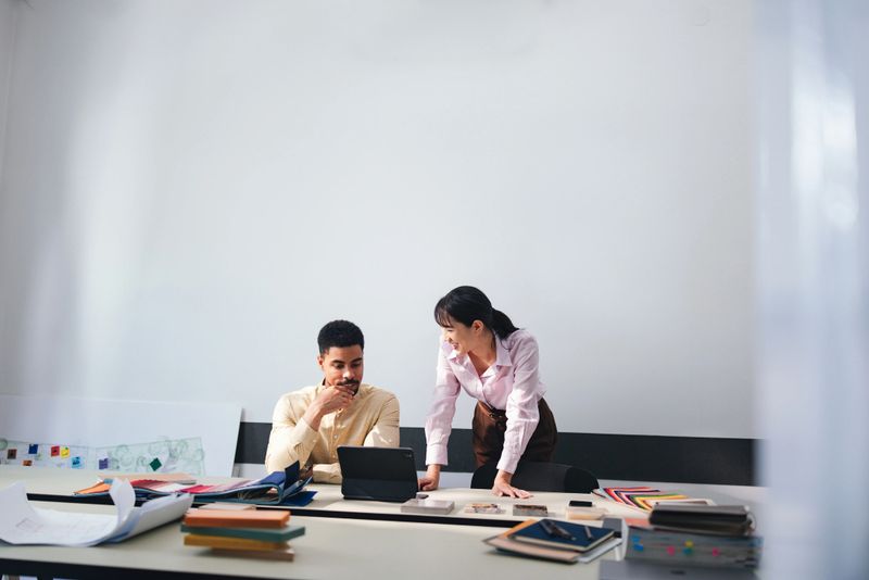 Two professionals collaborate in a bright, modern office. A man reviews content on a tablet while a woman leans over the desk, surrounded by color swatches and documents, signaling teamwork and design planning.