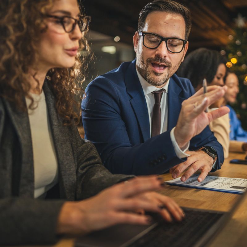 Business professionals engaging in a focused discussion at a table in the workplace, with a man explaining a concept to a woman typing on a laptop, fostering teamwork and shared learning
