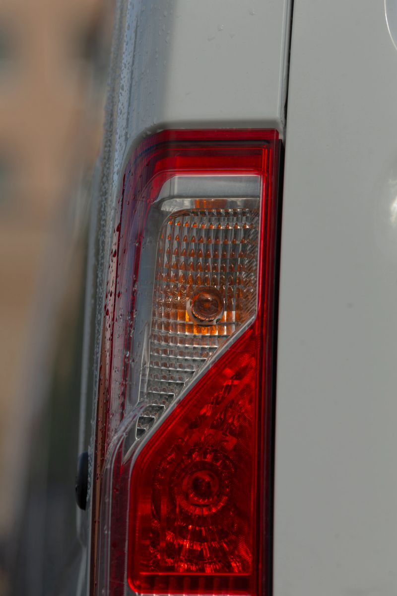 Vertical taillight wet at dusk on van, parking attendant prepping vehicle for nighttime delivery warm amber glow contrasts with cool wet body, droplets trail down lens with urban blur.