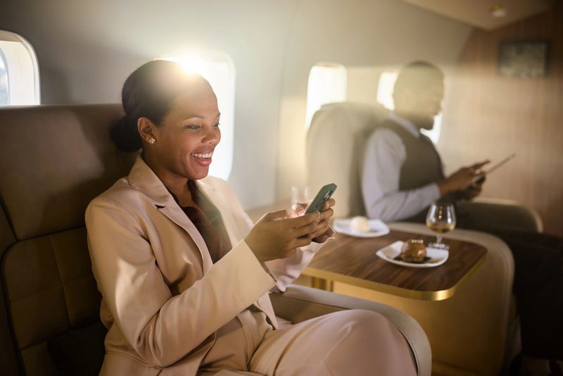Happy African American businesswoman text messaging on smart phone during travel by corporate jet. Her colleague is in the background.