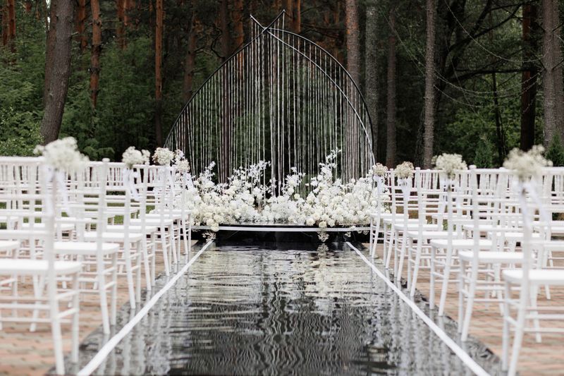An outdoor wedding ceremony setup with rows of white chairs and a beautiful floral arch surrounded by a forest. The elegant minimal decor soft white flowers and mirrored aisle create a romantic atmosphere perfect for themes of weddings celebrations romance and outdoor events.