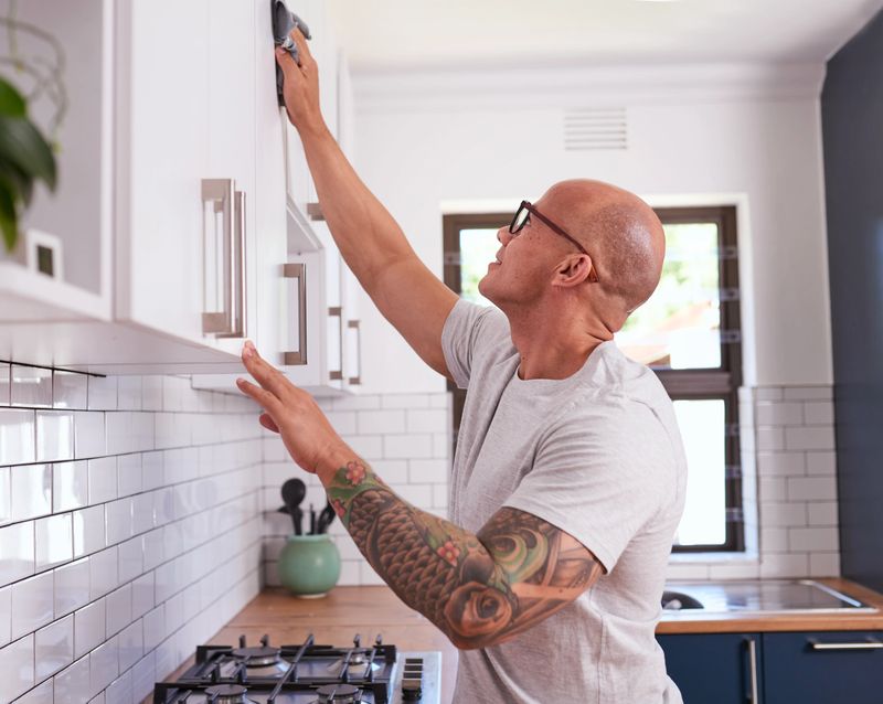 A bald, tattooed man wearing glasses cleans white kitchen cabinets, reaching upward with a cloth. Modern kitchen with subway tile, stove, and window letting in natural light, conveys home improvement and DIY effort.