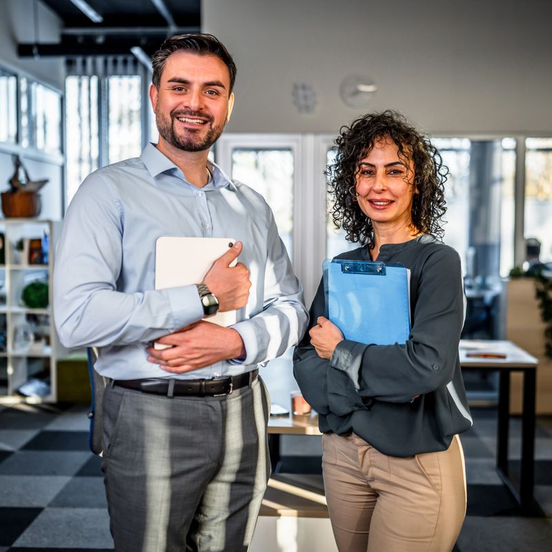 Two smiling multi-ethnic business colleagues standing confidently in a bright modern office, holding a tablet and clipboard