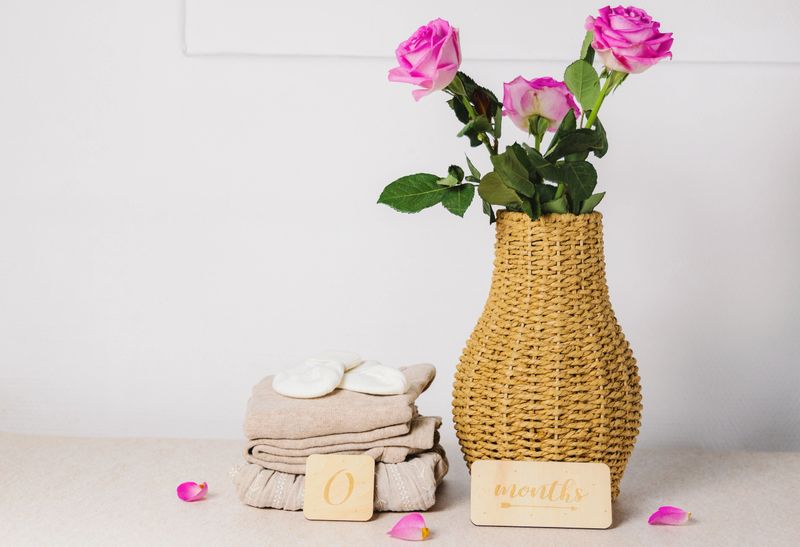 Beautiful beige knitted baby clothes are stacked with booties and wooden 0-month signs, next to a wicker vase with pink roses and scattered petals on a table against a white wall, close-up side view. Concept: Baby clothes, child's height.