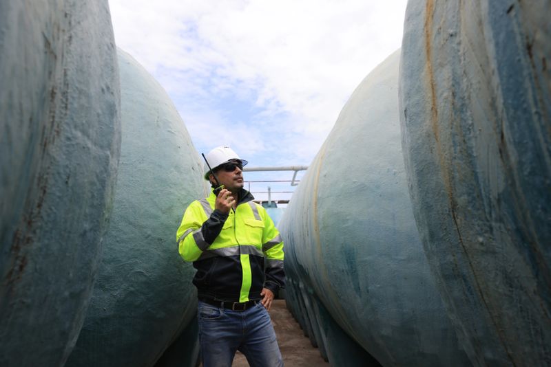 An industrial worker in a hard hat and fluorescent jacket inspects large water tanks while holding a communication device, Water storage and supply system