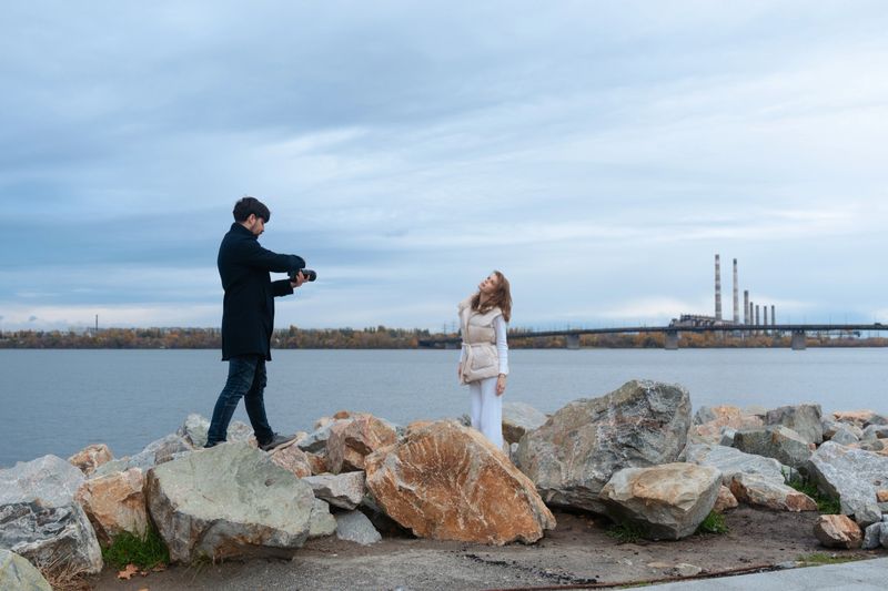 A photographer takes a picture of a woman standing on a rocky shoreline with a lake and bridge in the background.