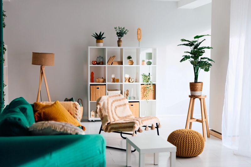 Contemporary cozy bohemian living room with teal sofa, patterned pillows, wooden lamp, knit pouf, white shelving and abundant plants for a relaxed, stylish home vibe