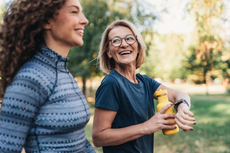 Two women, friends enjoying an outdoor workout or run together in a park. One woman is smiling brightly as she checks her smartwatch, possibly tracking time, distance, or heart rate. They both walking and talking friendly, appear relaxed and happy, surrounded by lush greenery and warm sunlight, creating a positive and energetic atmosphere.