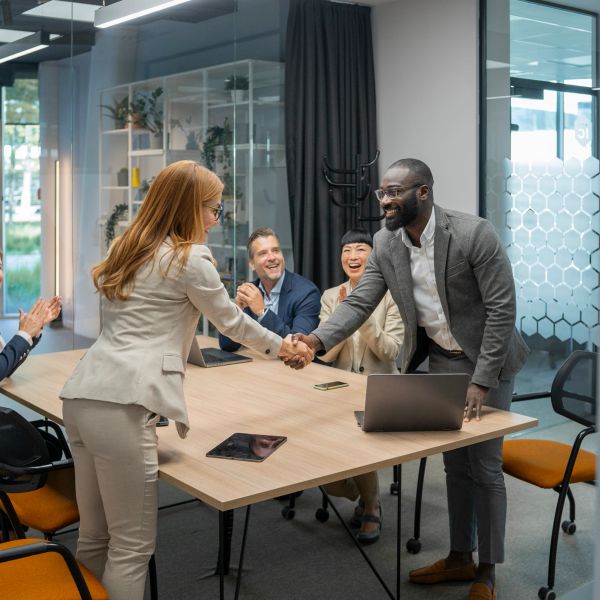 Two businesspeople shaking hands in a meeting room with colleagues applauding.