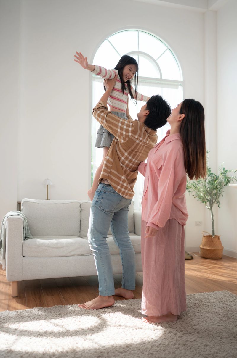A father lifting his daughter playfully in a bright living room while the mother watches with a smile