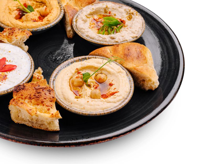 A black plate holds various types of hummus alongside warm pita bread. The creamy dips are garnished with spices and herbs, showcasing a colorful and inviting presentation.