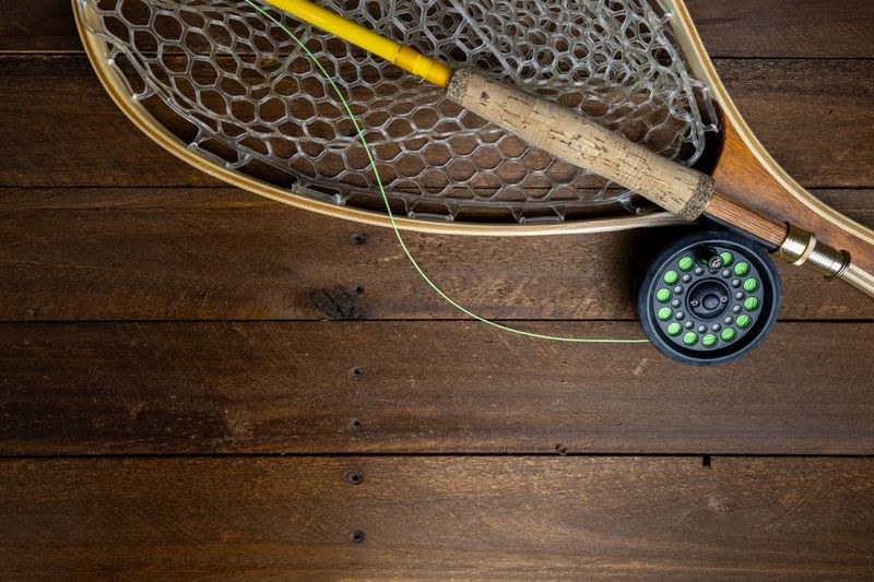Overhead flat lay of essential fly fishing gear: net, cork-grip rod handle, and reel with bright green line, resting on rustic dark wood planks.