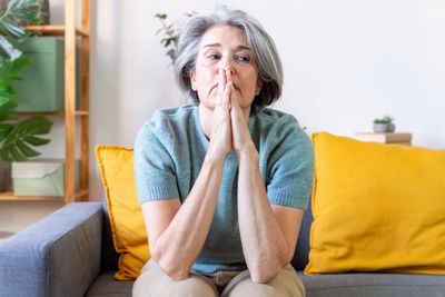 Pensive older woman sitting on couch with hands clasped.