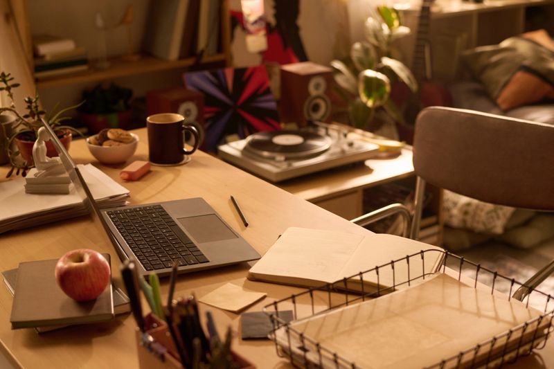 Home office desk featuring open laptop, notebook, apple, stationery, vinyl record player and bookshelves in background, workspace arranged for remote work or creative projects