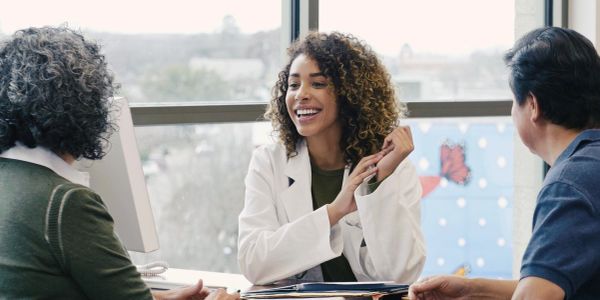 A cheerful woman in a white coat talking to two people at a desk.