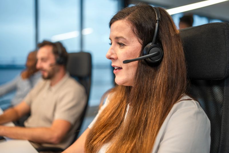 Mid adult Caucasian female professional wearing a headset and communicating in an open plan office setting, focused on managing tasks and collaborating with colleagues.
