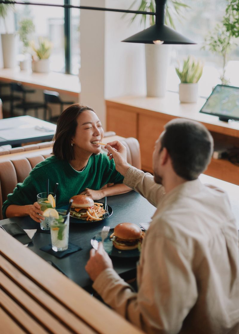 A young, adult married couple share a playful moment at a cozy restaurant table, feeding each other and smiling. They enjoy burgers, fries, and drinks, conveying warmth, affection, and romantic date vibes.