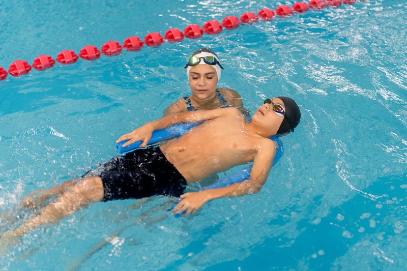 Young boy learning to float on his back in a swimming pool, wearing googles and swim cap, an instructor supervising