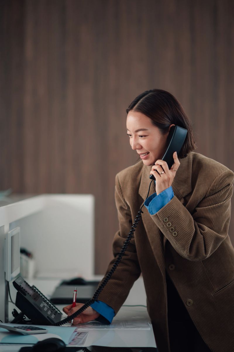 Smiling businesswoman talking on a landline phone and providing customer service while writing at a reception desk