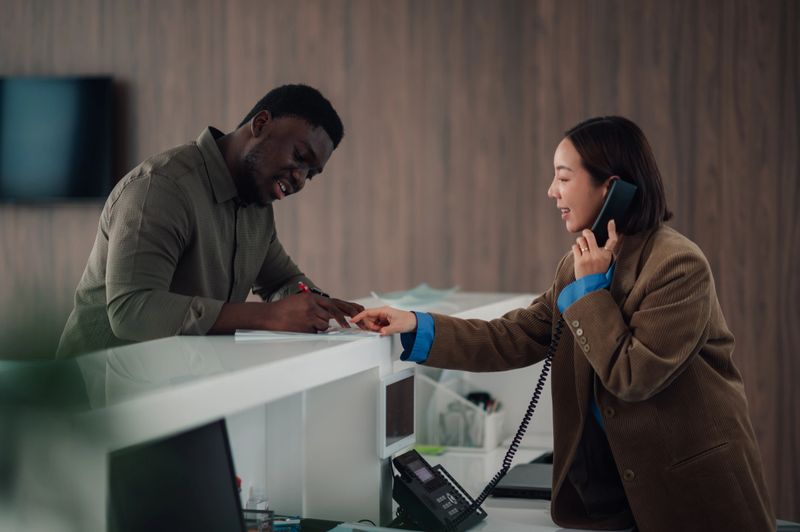 Man signing documents at a reception desk while a woman talks on the phone, providing customer service in an office