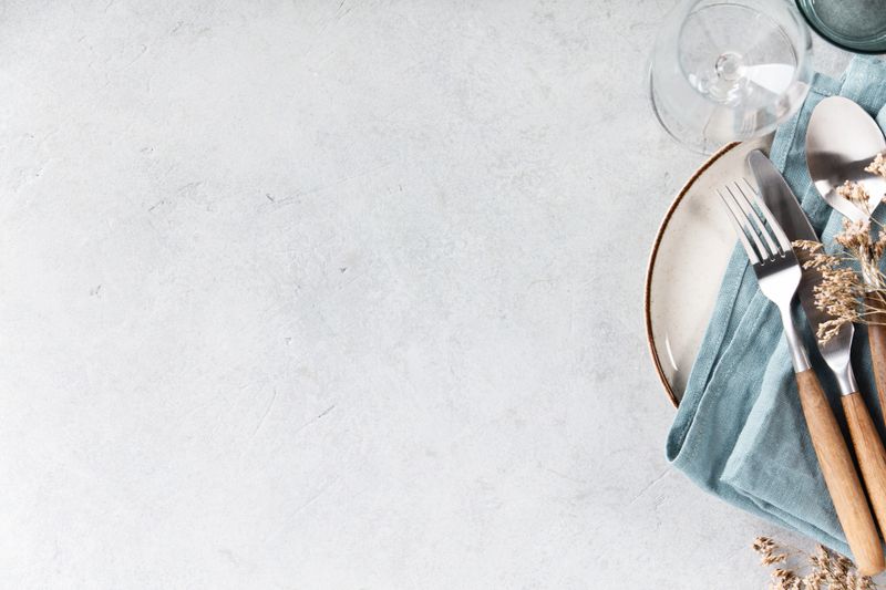 Top view of a minimal and elegant table setting featuring a ceramic plate with blue linen napkin, wooden-handled cutlery, and glassware on a light textured background. Dried flowers add a rustic touch. Perfect for food, lifestyle, or hospitality themes.