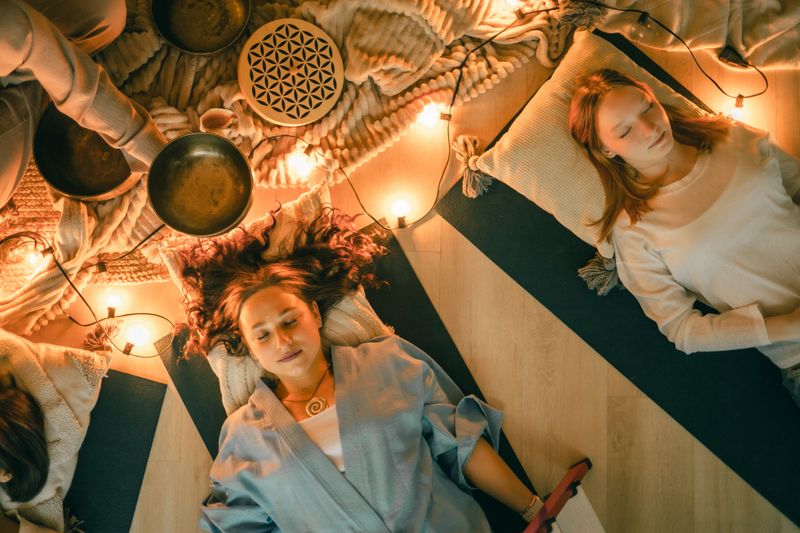 Women lying on mats with eyes closed during a calming sound healing session surrounded by warm lights, singing bowls, soft textiles, and peaceful energy for deep relaxation and mindfulness.