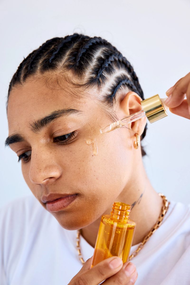 Closeup shot of a young person looking down while applying facial serum with a dropper in a studio, showing flawless skin and braided black hair.