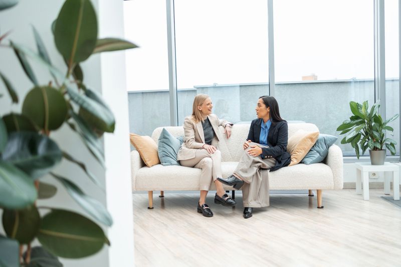 Two professional businesswomen on a sofa in a modern office lounge, engaged in a focused discussion about strategy and collaboration