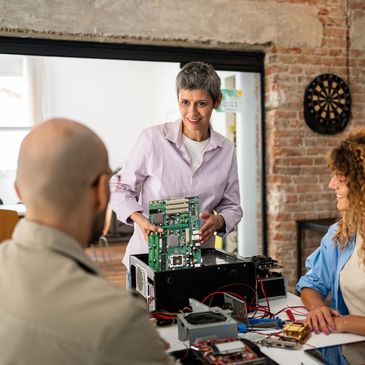 A woman explains a computer motherboard to two attentive colleagues in a modern workspace.