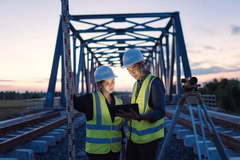 Two civil engineers examining project data on railway bridge construction site using measurement tools for accurate planning. Surveyor team measuring position on railway with level tool and tripod.