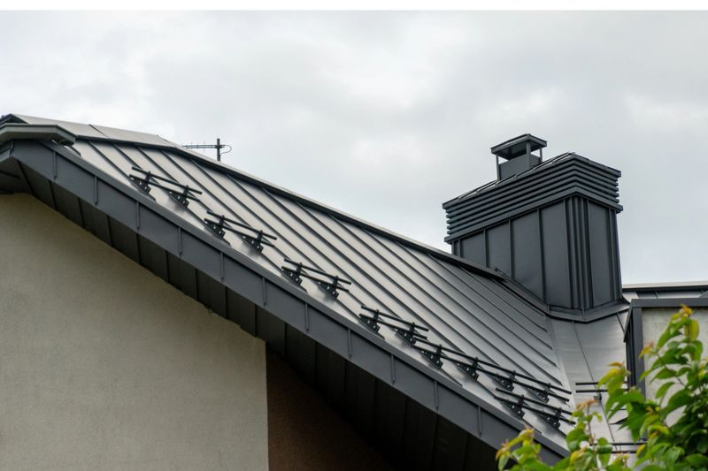A close view of a roof with metal panels and a chimney against a cloudy sky in a residential neighborhood.