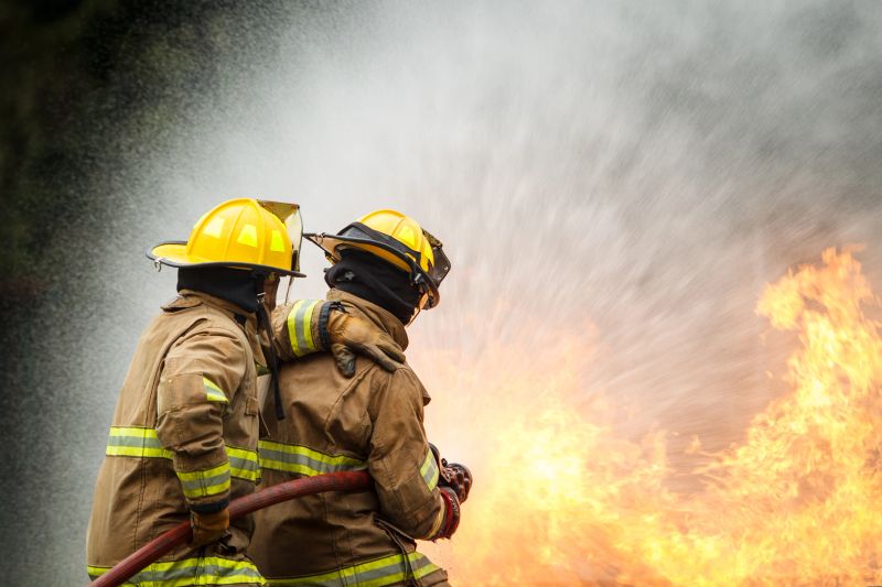 Firefighter in protective gear uses high-pressure hose to spray water toward intense flames during live fire operation. Emergency response, fire suppression skills, and professional safety training.