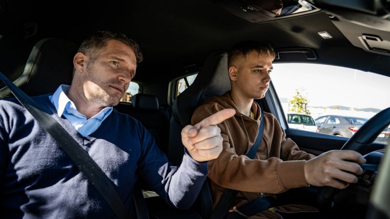 A focused driving instructor in a blue sweater guides a young learner in a brown hoodie, emphasizing safety and control. Sunlight highlights the learning environment.