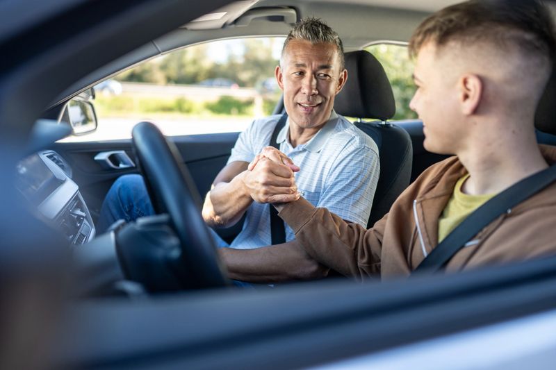 Two men in a car; the driver, a middle-aged man, smiles as he shakes hands with a younger passenger. The setting suggests a driving lesson, emphasizing guidance and learning. The instructor's supportive demeanor and the student's attentive posture highlight the educational context.