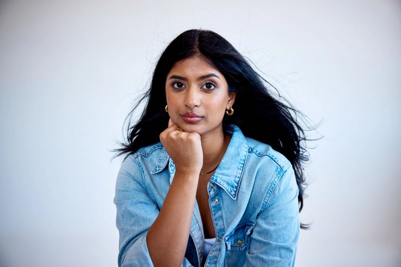 Closeup portrait of a flawless young woman with flowing black hair and glowing skin resting her chin on her hand in a studio, looking at camera with confidence.