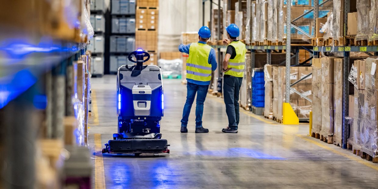 Two workers discuss near an autonomous floor cleaning robot in a warehouse.