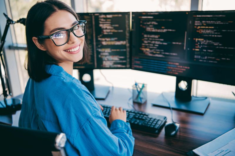 A focused young professional works at a desk with multiple monitors displaying code, keyboard and mouse in a bright loft office environment