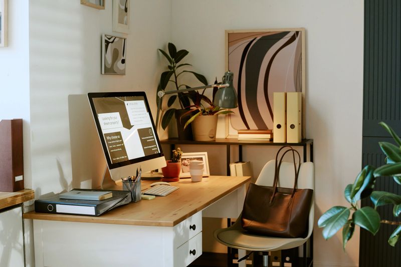 Modern home office workspace featuring desktop computer displaying charts and documents, organized desk with office supplies, indoor plants and bookshelves in background