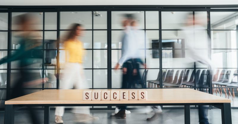 Modern office environment with several people walking past in a blur, creating a sense of motion and busyness. In the foreground, on a wooden table, wooden letter blocks spell out the word “SUCCESS.” The contrast between the stillness of the word and the fast-paced movement of the people around it suggests themes of productivity, teamwork, and progress. The glass-panel wall in the background gives the scene a clean, contemporary feel.