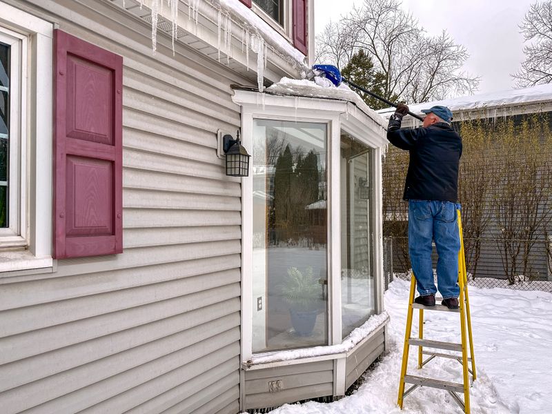 Senior man removing snow and ice from roof of bay window