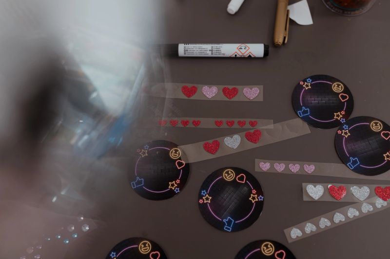 Glitter heart stickers placed near neon-style round labels with Cyrillic phrases arranged across craft desk during kids decoration activity featuring pens, tape, scattered materials.
